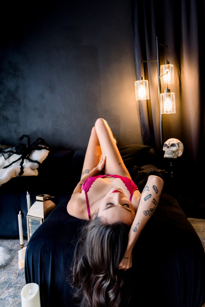 Dark and moody boudoir portrait in South Orange County, woman in magenta lace draped over a black velvet sofa beside a skull 